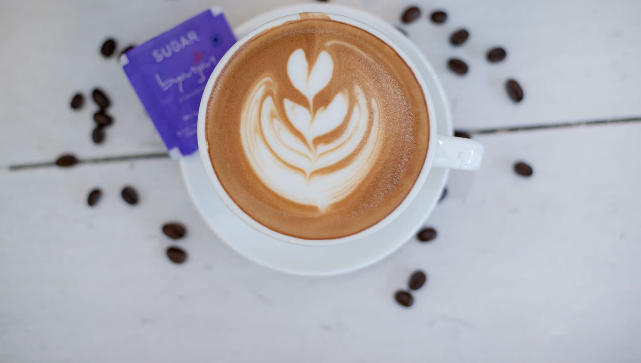 Cappuccino with leaf latte art, a Byanjan sugar sachet and coffee beans on white wood.