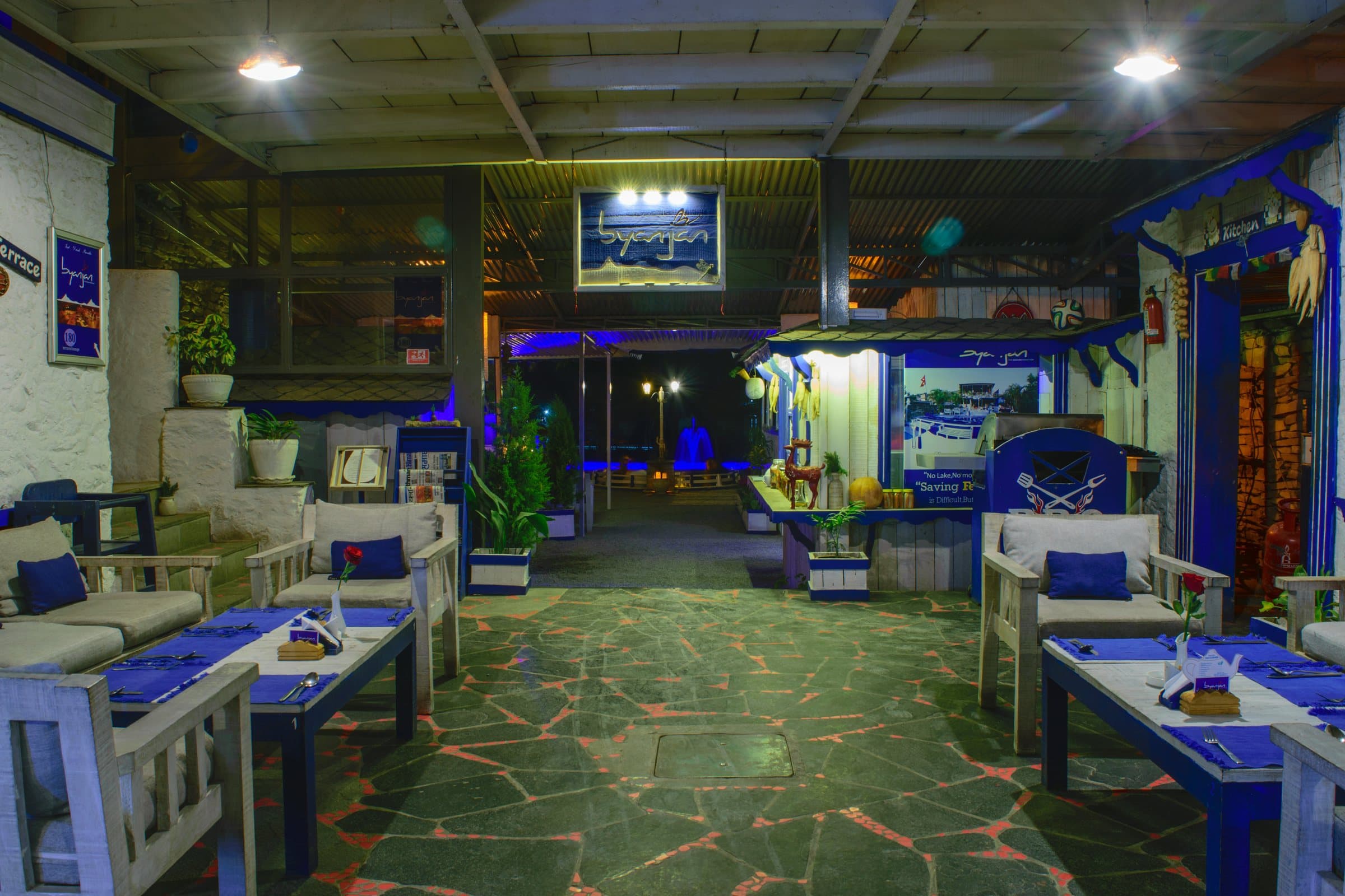 The dining room at night: stone floor, blue-lit acrylic panels and white chairs at blue tables.
