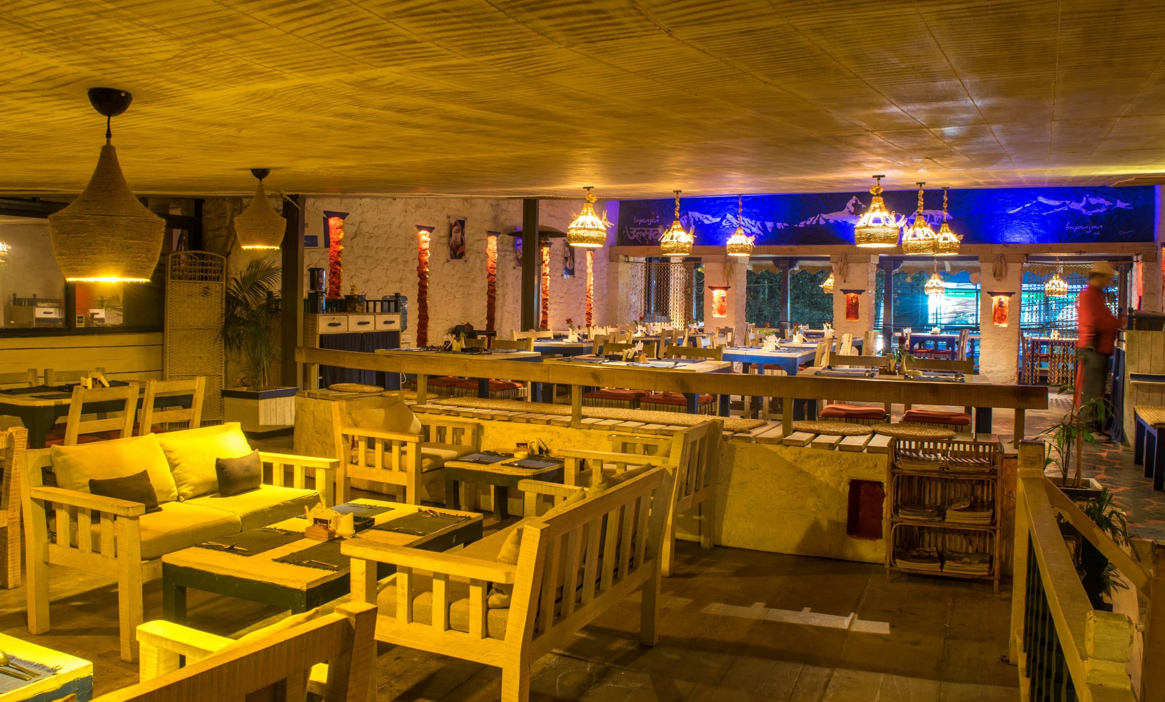 The dining room in lamplight: wooden benches and tables, woven lanterns overhead, blue light along the far wall.